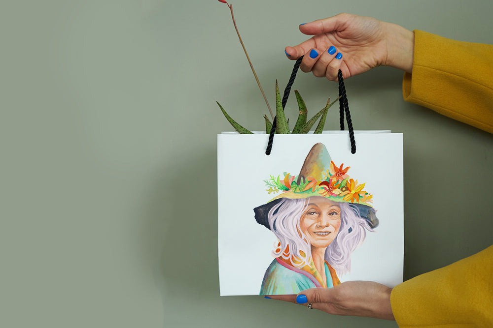 A person holds a white paper shopping bag with black handles against a muted sage green background. The bag features a colorful printed illustration of a smiling witch with a flower-crowned hat.