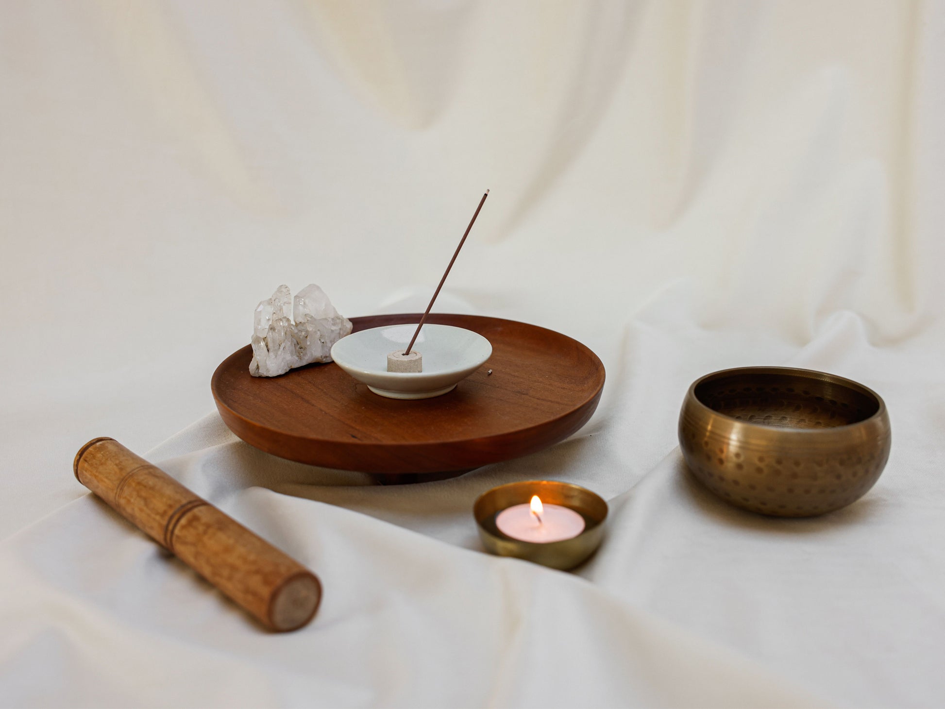 A styled arrangement of meditation and yoga props on draped white fabric, including a wooden tray with a small white dish holding an incense stick, a white crystal cluster, a brass singing bowl, a wooden mallet, and a small lit candle.