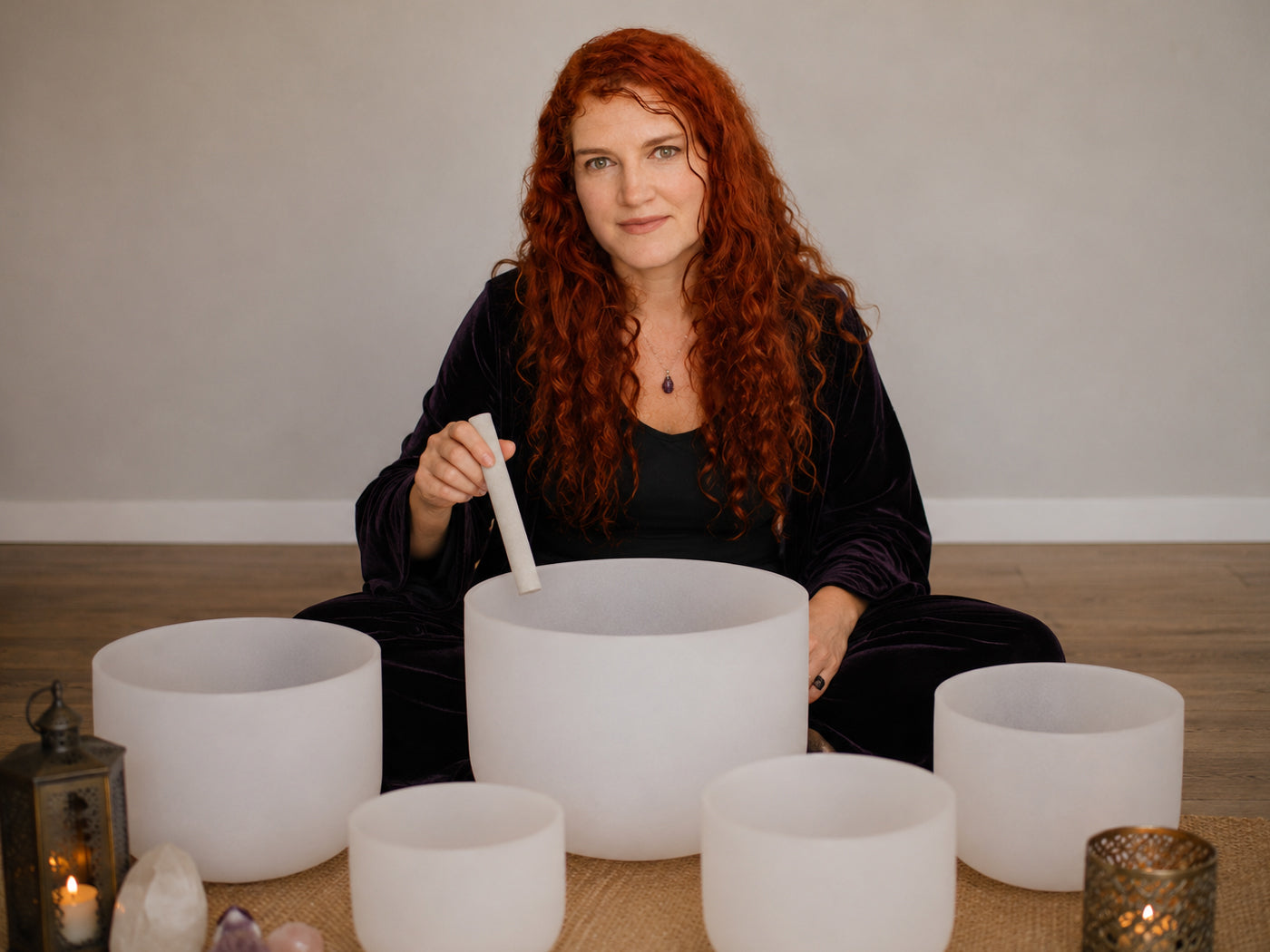 A woman with long red hair sits cross-legged on a wooden floor in front of a large white singing bowl set, holding a white mallet. The set includes multiple white crystal singing bowls, a black velvet robe, and decorative candles and crystals on the floor.